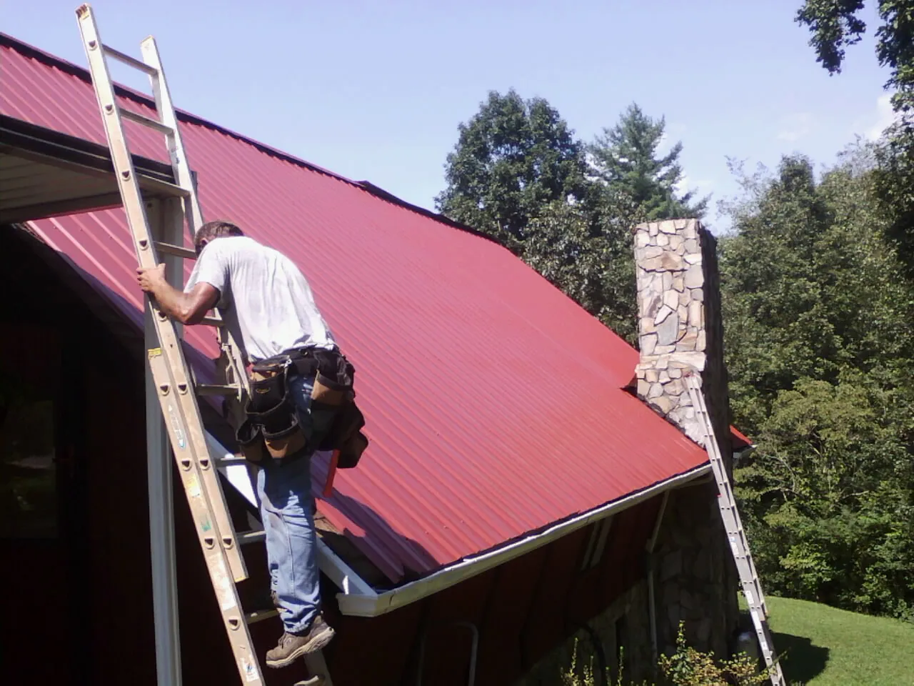 Roofer on a ladder beside a red metal roof and stone chimney — Soffit & Fascia Repair by Roofing Brothers in Mocksville